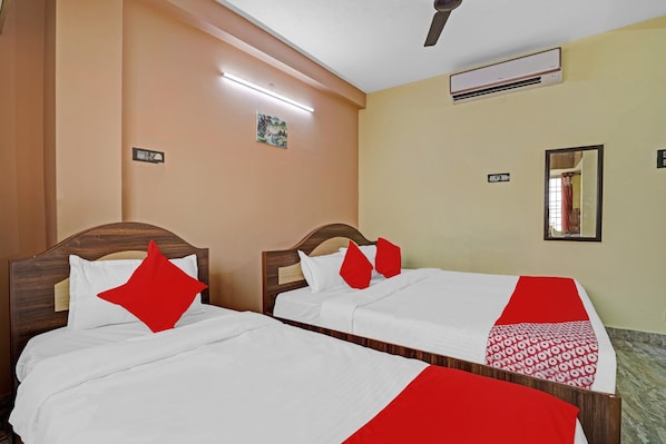 A hotel room with two beds, each with white bedding and red accents, a ceiling fan, an air conditioning unit, and a small painting on the wall.