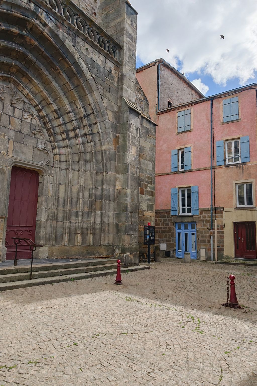 A cobblestone courtyard with a stone building featuring a red door and a pink building with blue shutters.