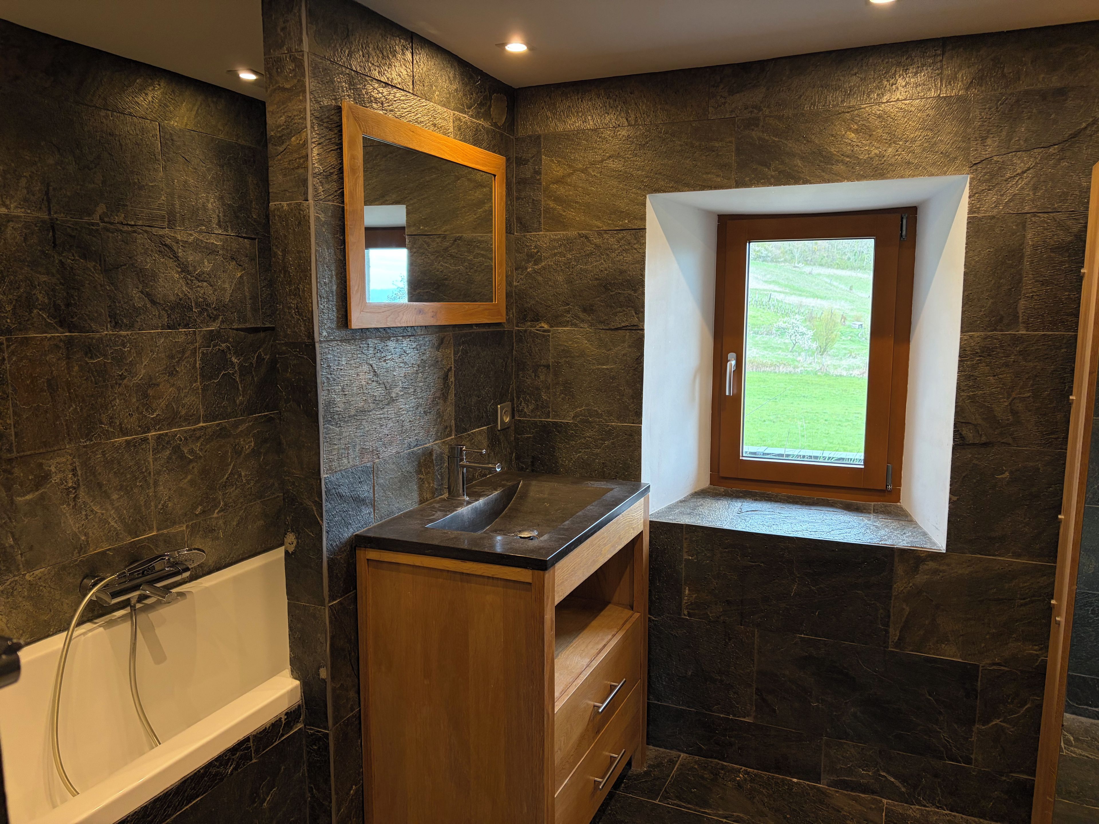 A bathroom with a bathtub, a wooden vanity, a mirror, and a window with a view of greenery.