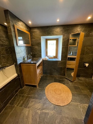 A bathroom with a stone wall, a wooden vanity, a bathtub, a window, and a mirror. - Basel Landskron Haus (LEYMEN)