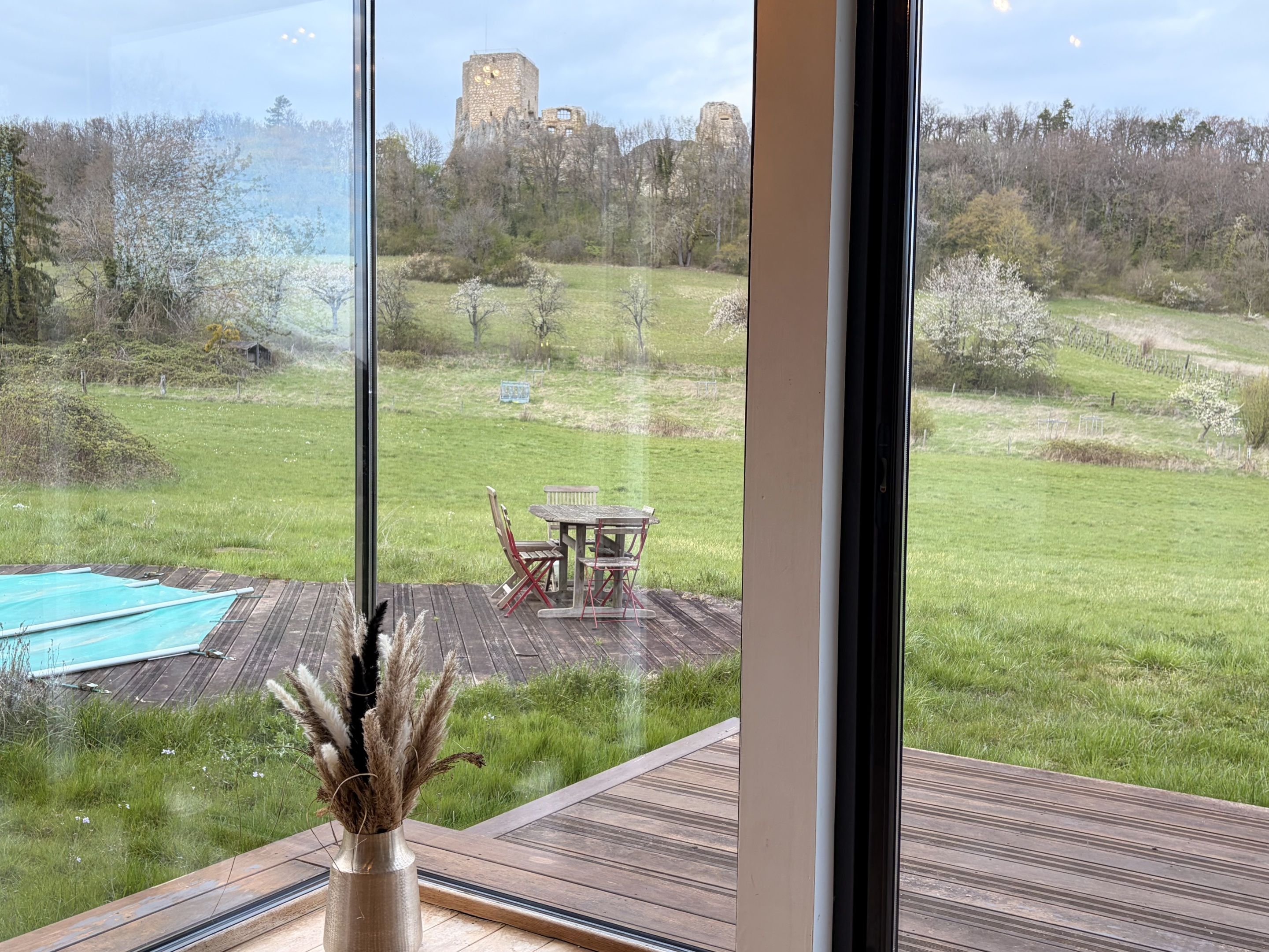 A view from a room with a large window overlooking a grassy area with a wooden deck, a table, and chairs.