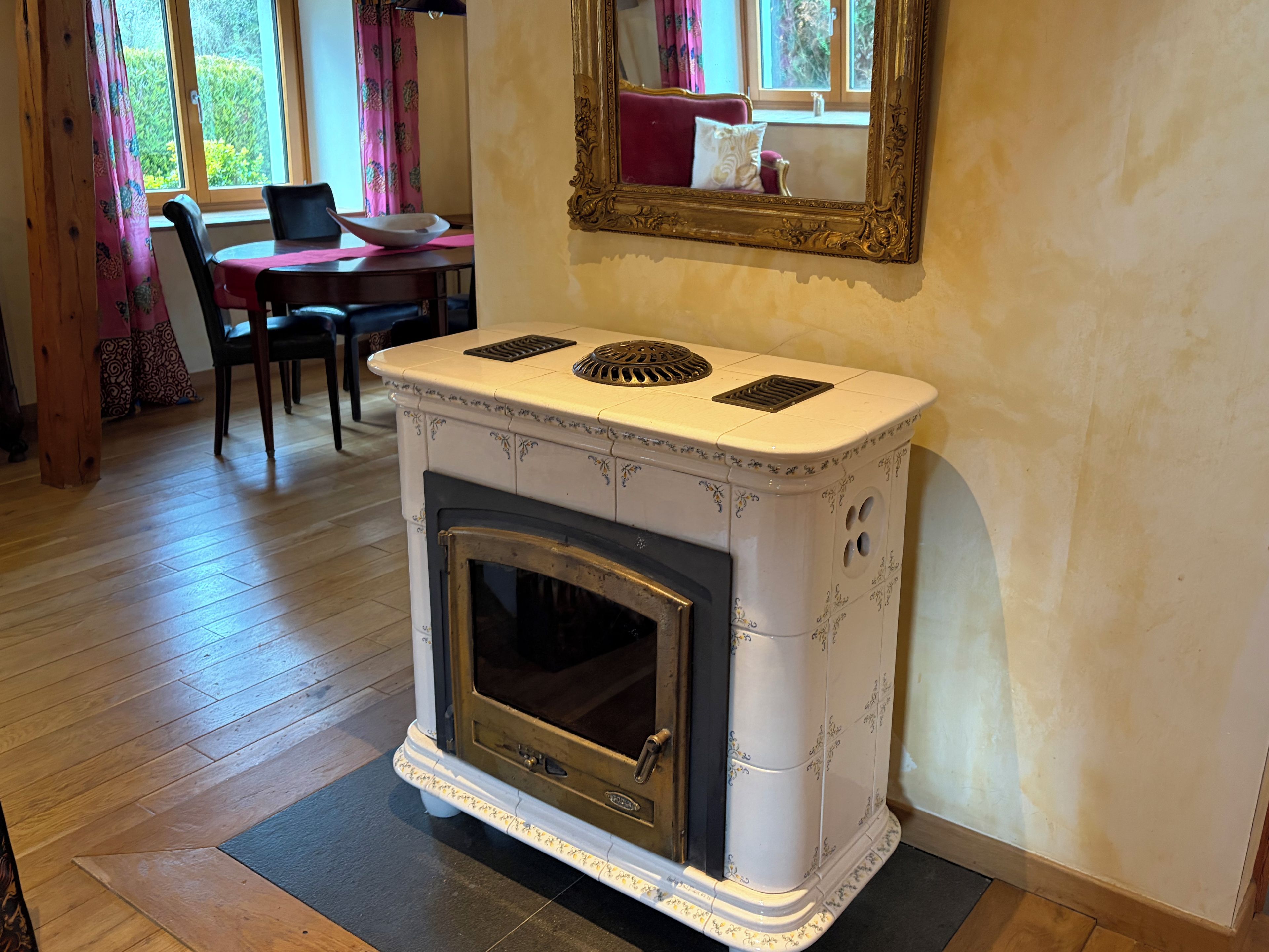 A room with a white tiled stove, a dining table with chairs, and a window with curtains.