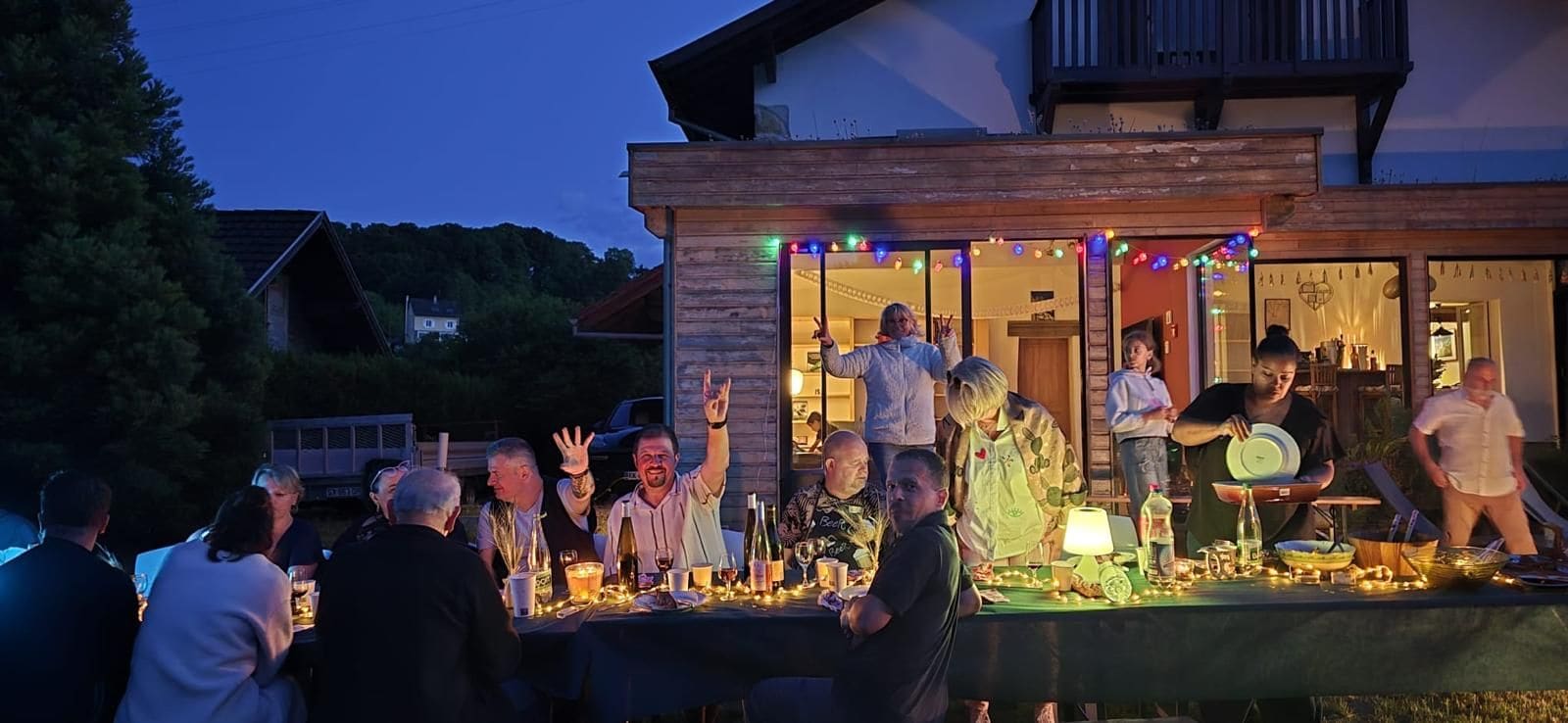 An outdoor gathering with people seated at tables, some standing, and a building with a balcony in the background.