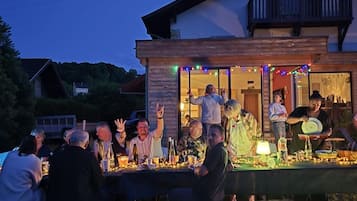 An outdoor gathering with people seated at tables, some standing, and a building with a balcony in the background.