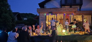 An outdoor gathering with people seated at tables, some standing, and a building with a balcony in the background. - Basel Landskron Haus (LEYMEN)