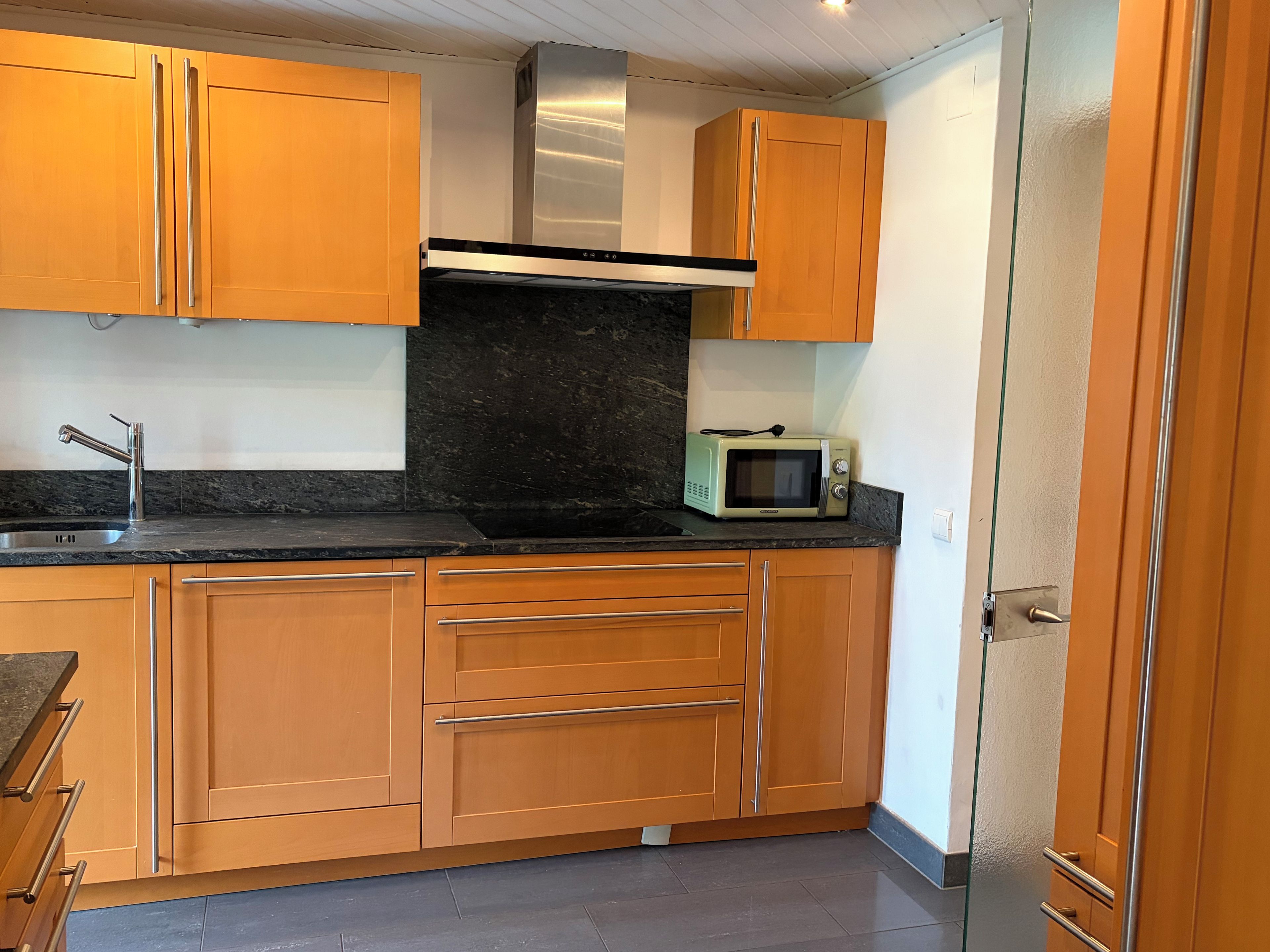 Cocina compacta con armarios de madera, campana extractora de acero inoxidable, microondas y pileta con grifo moderno.