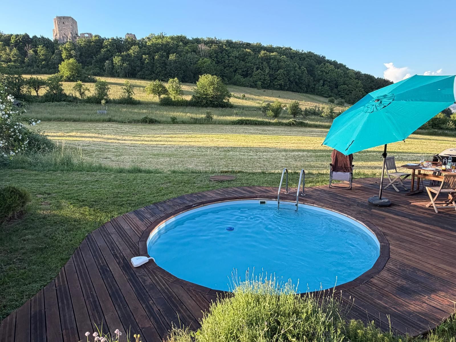 A swimming pool with a wooden deck, a table with chairs, and an umbrella.