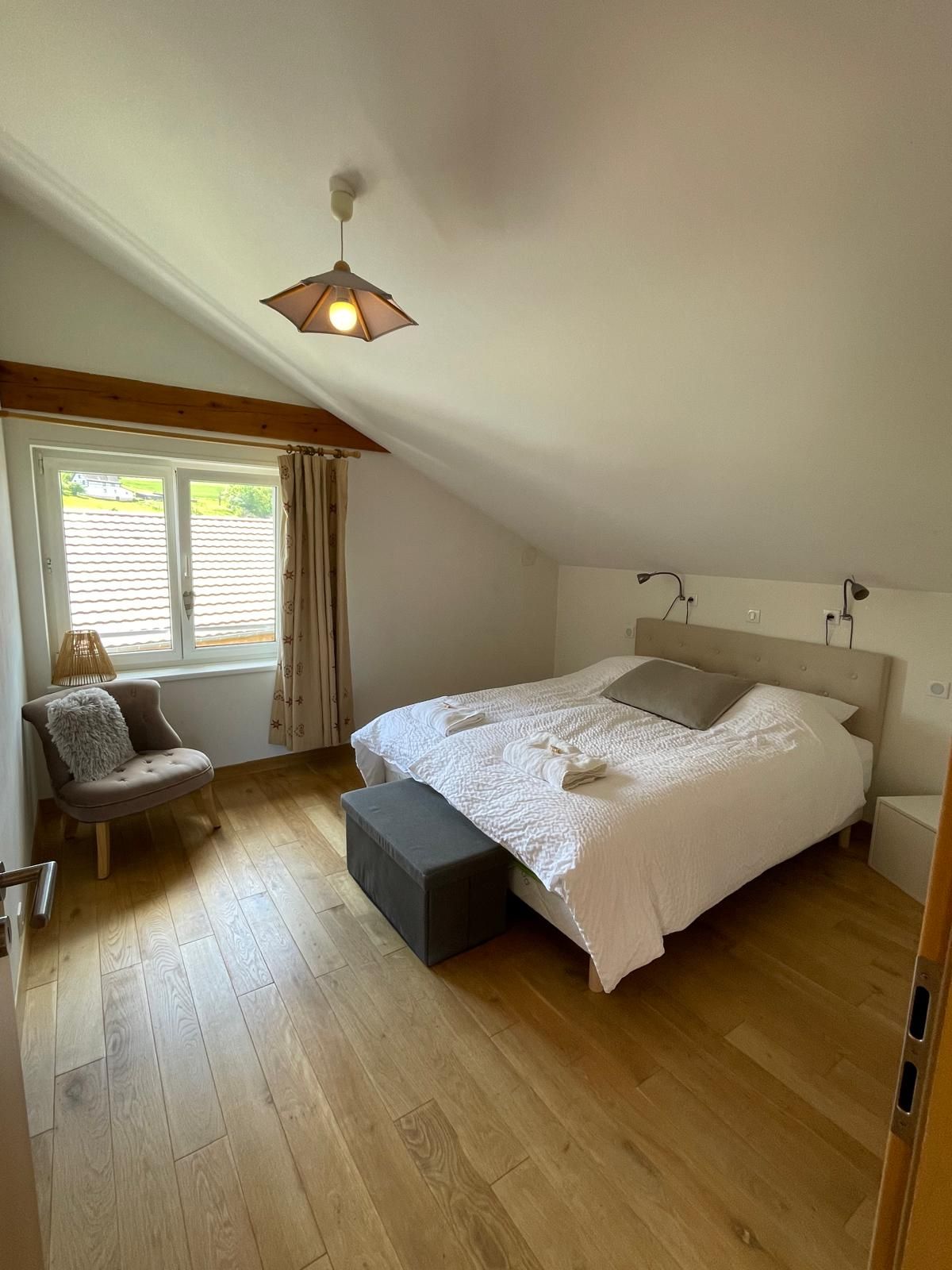 A bedroom with a large bed, a chair, a window with curtains, and a sloped ceiling.