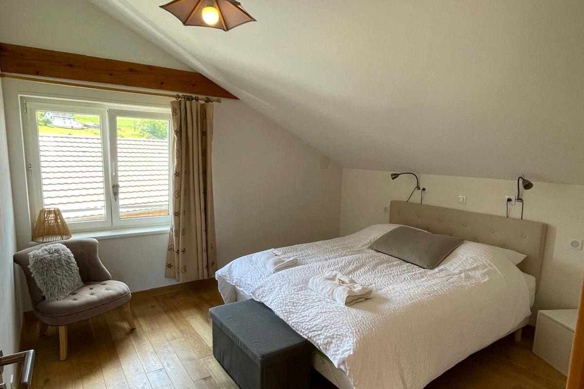 A bedroom with a large bed, a chair, a window with curtains, and a sloped ceiling.