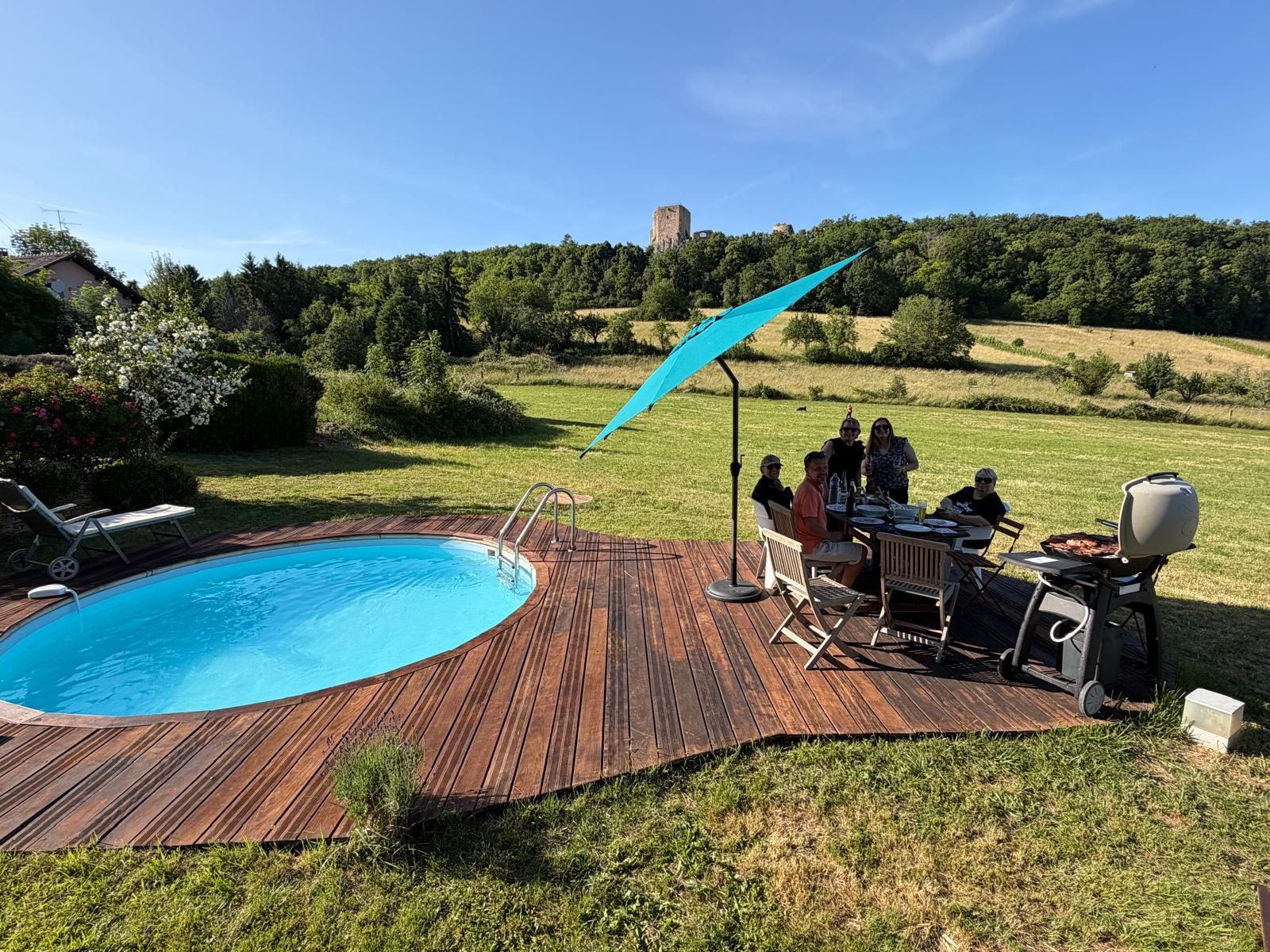A poolside area with a wooden deck, a barbecue grill, and a group of people dining.