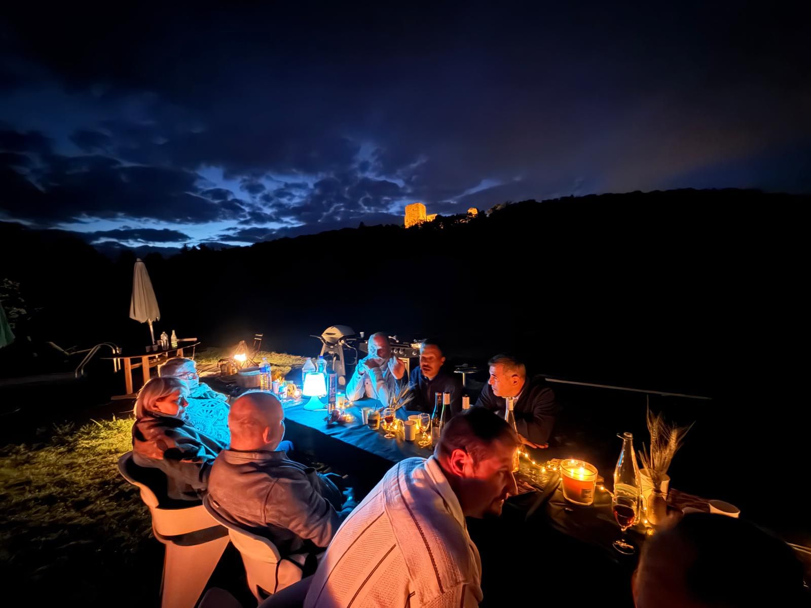 A group of people dining outdoors at night with a table set for a meal, surrounded by a scenic view of a hill and a building in the distance.