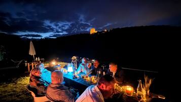 A group of people dining outdoors at night with a table set for a meal, surrounded by a scenic view of a hill and a building in the distance.