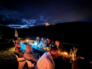 A group of people dining outdoors at night with a table set for a meal, surrounded by a scenic view of a hill and a building in the distance. - Basel Landskron Haus (LEYMEN)