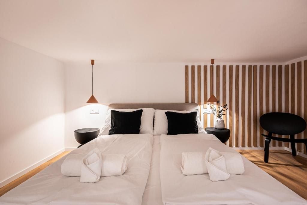 Uma cama de casal com roupa de cama branca, duas almofadas pretas, uma cabeceira de madeira, uma mesa de cabeceira preta com uma luminária, uma cadeira preta e uma parede listrada.