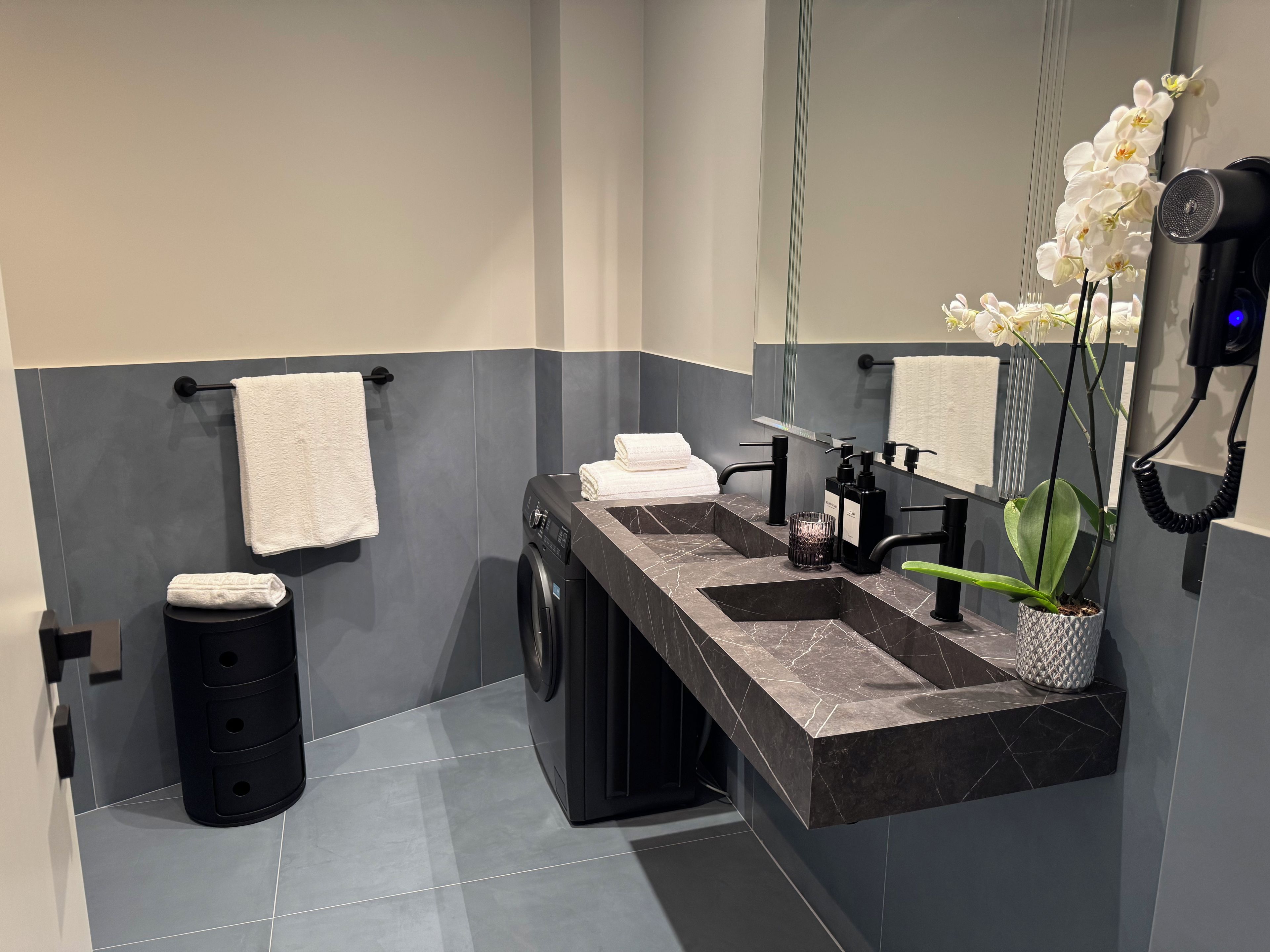 Une salle de bain moderne équipée d’un lave-linge intégré, d’un lavabo avec un plan de travail en marbre, d’un miroir et d’une orchidée en pot.