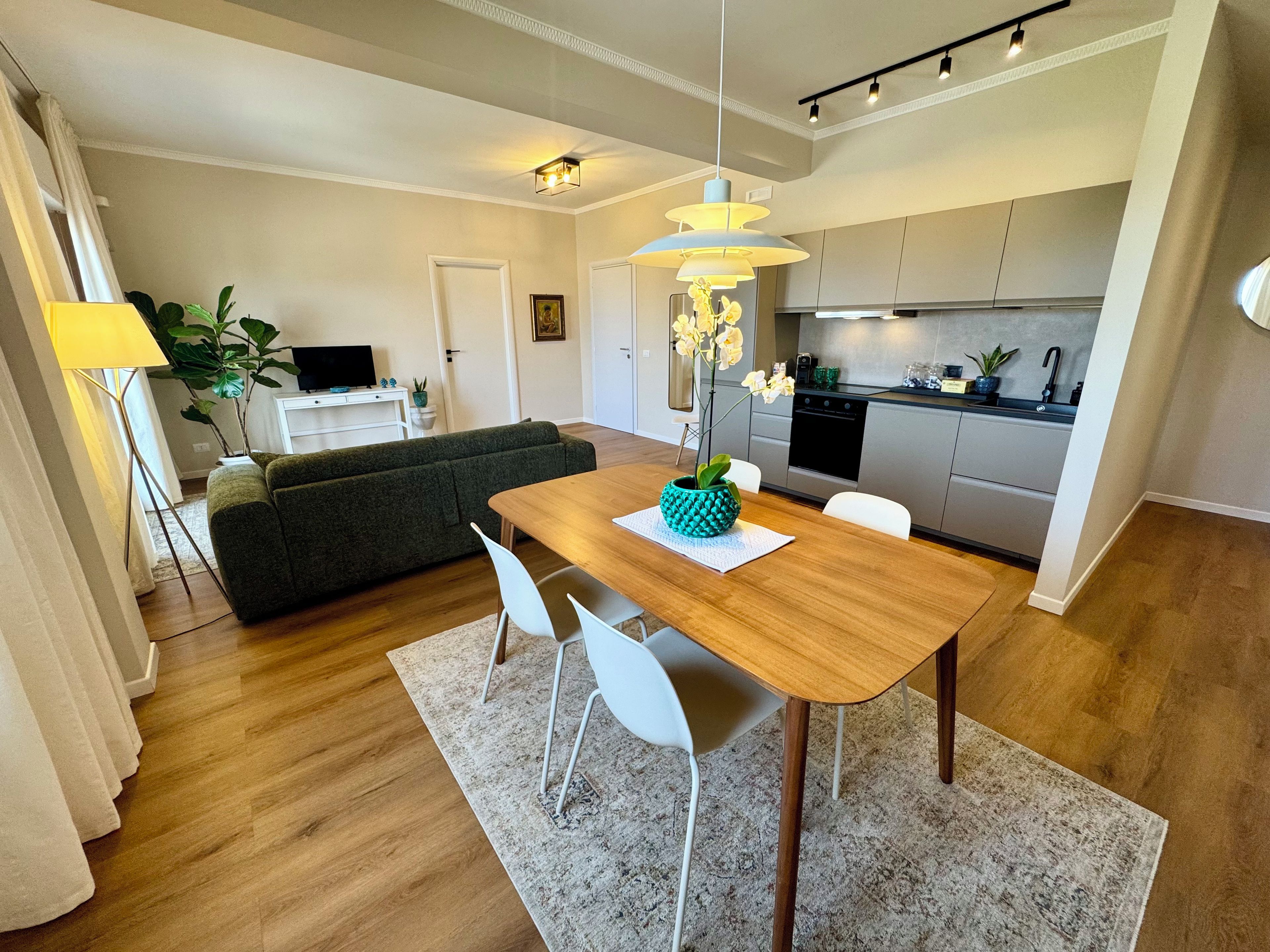 Une cuisine moderne dotée d’un four encastré, un coin repas avec une table en bois et des chaises blanches, et un canapé gris en arrière-plan.