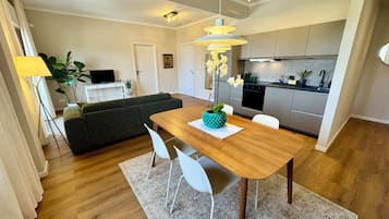 Une cuisine moderne dotée d’un four encastré, un coin repas avec une table en bois et des chaises blanches, et un canapé gris en arrière-plan.