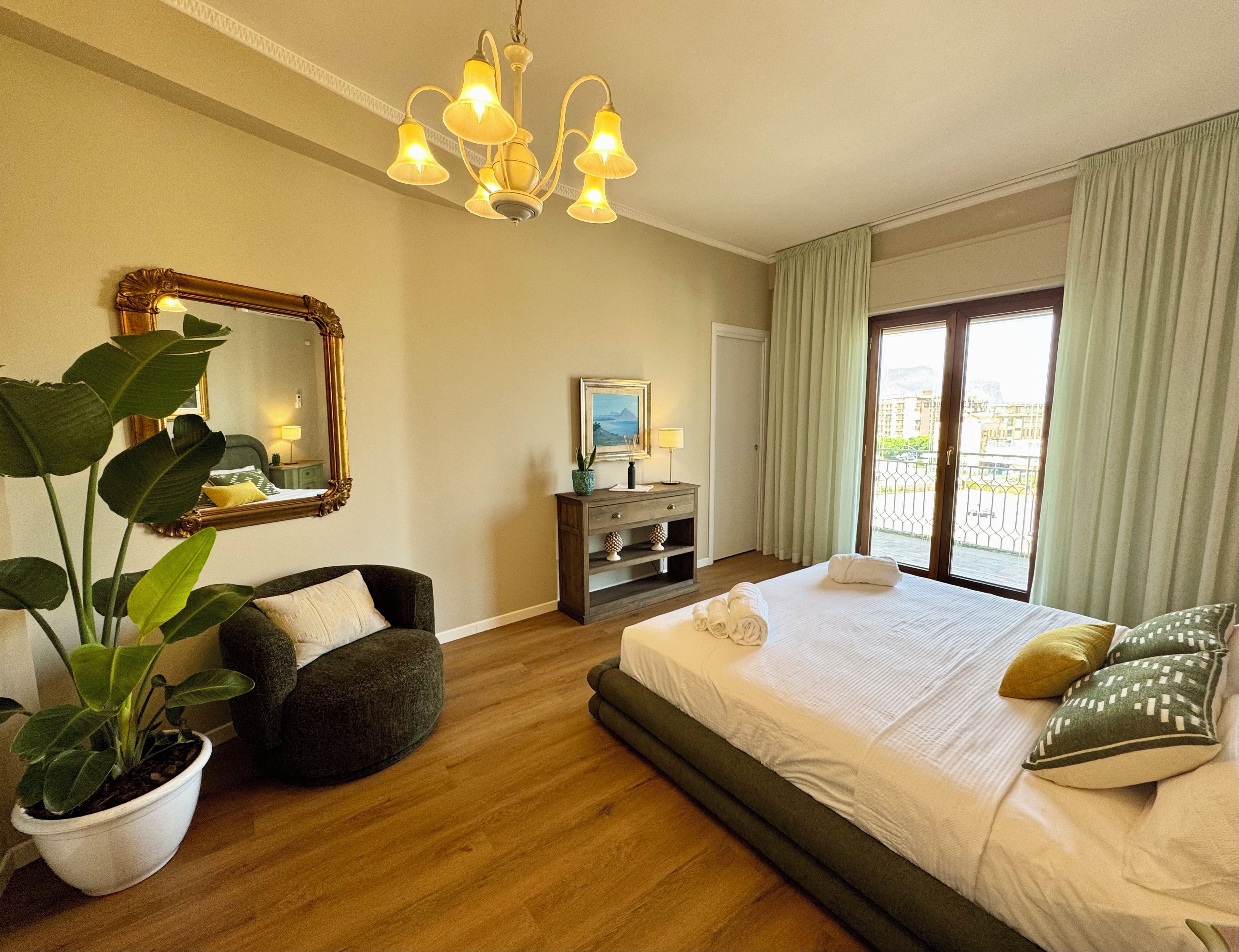 Une chambre d’hôtel avec un grand lit, un coin salon, un bureau avec une lampe, un grand miroir et une plante en pot.