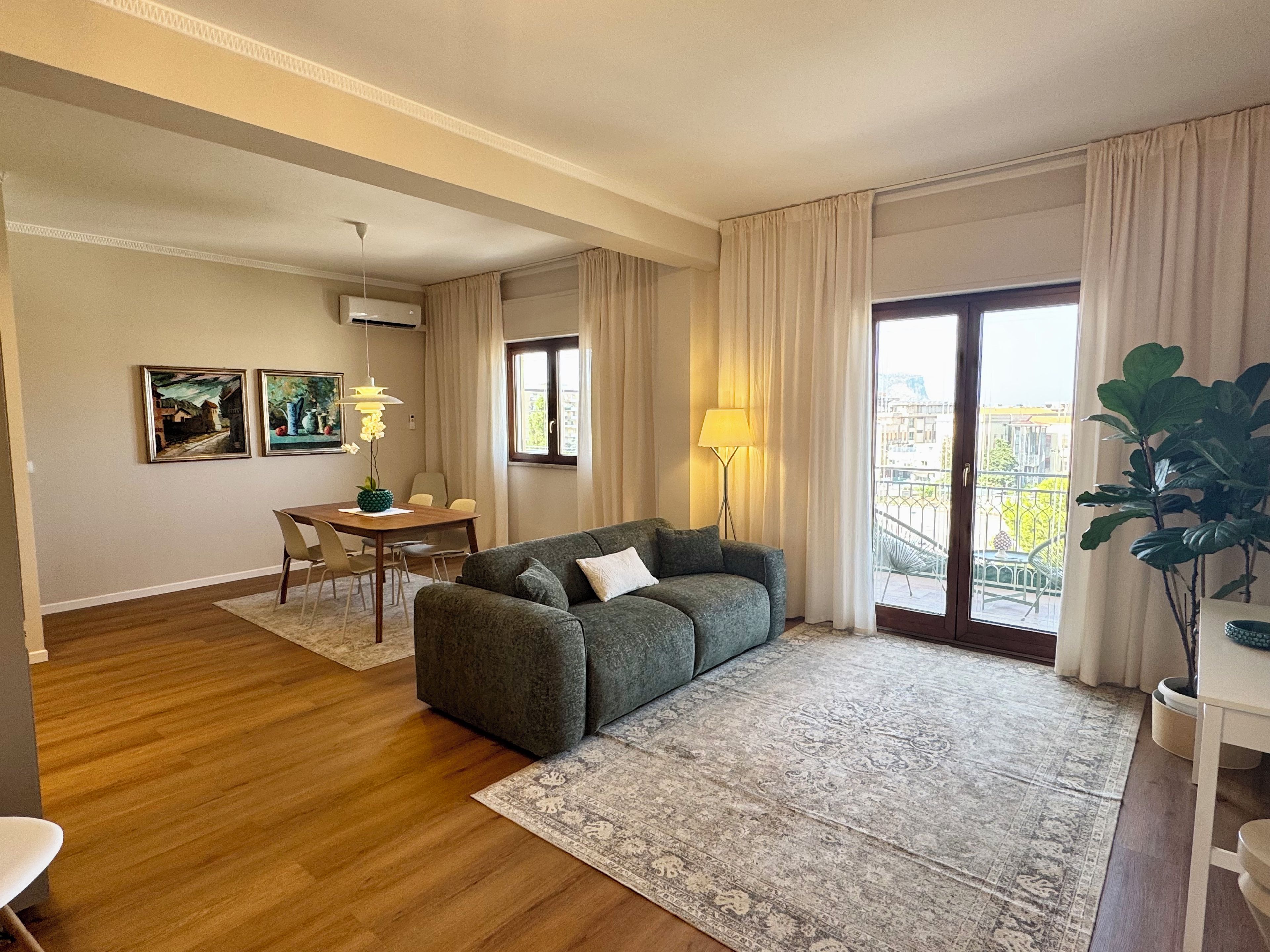 A modern living room with a sofa, dining table, and chairs, a large window with curtains, and a view of the city.