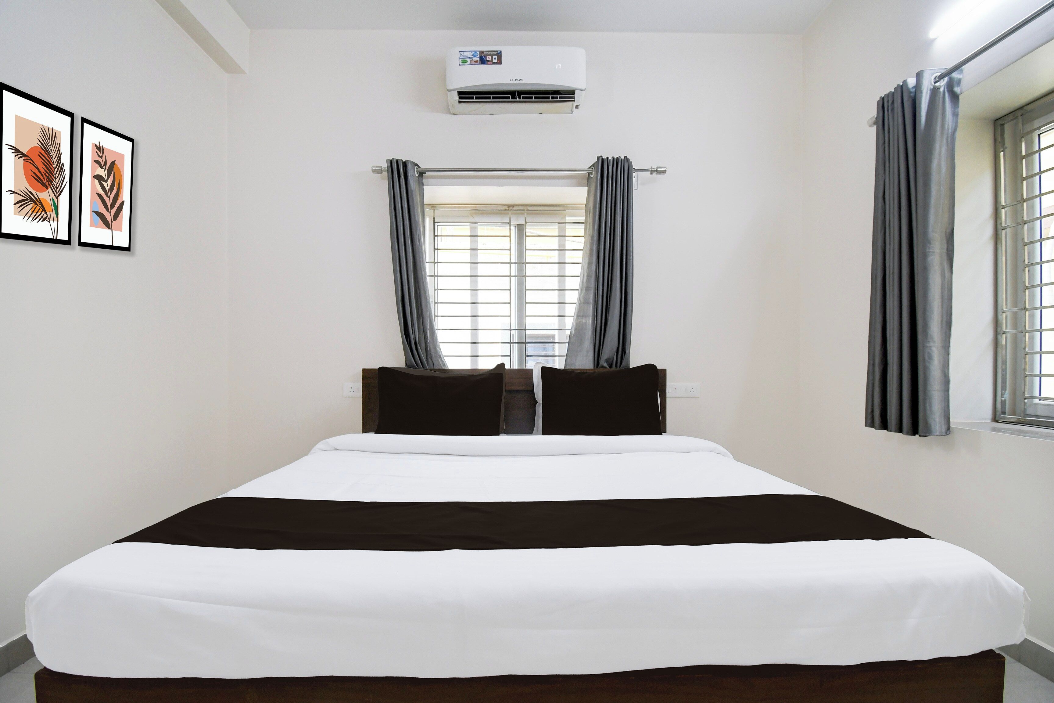 Une chambre d’hôtel comprenant un lit, un appareil de climatisation, une fenêtre avec des rideaux et deux œuvres d’art encadrées accrochées au mur.