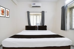 Habitación de hotel con cama, unidad de aire acondicionado, ventana con cortinas y dos obras de arte enmarcadas en la pared.