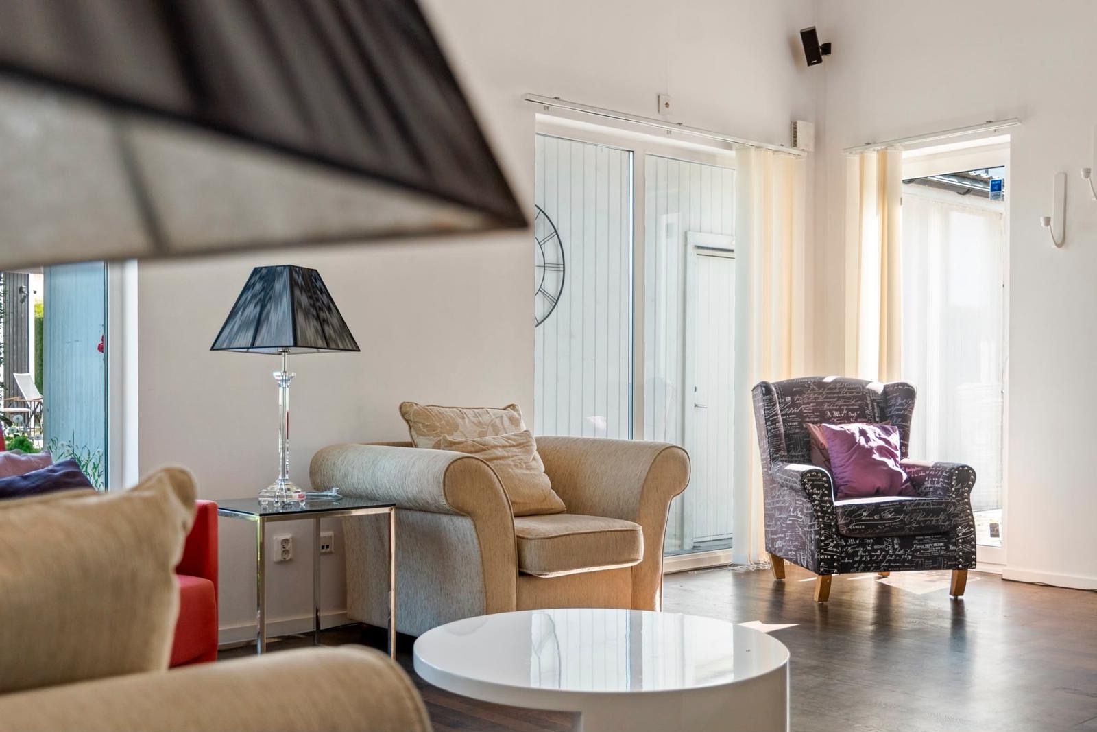 A modern living room with a sofa, armchair, and a table. There is a lamp on a side table, and a sliding glass door leading to an outdoor area.
