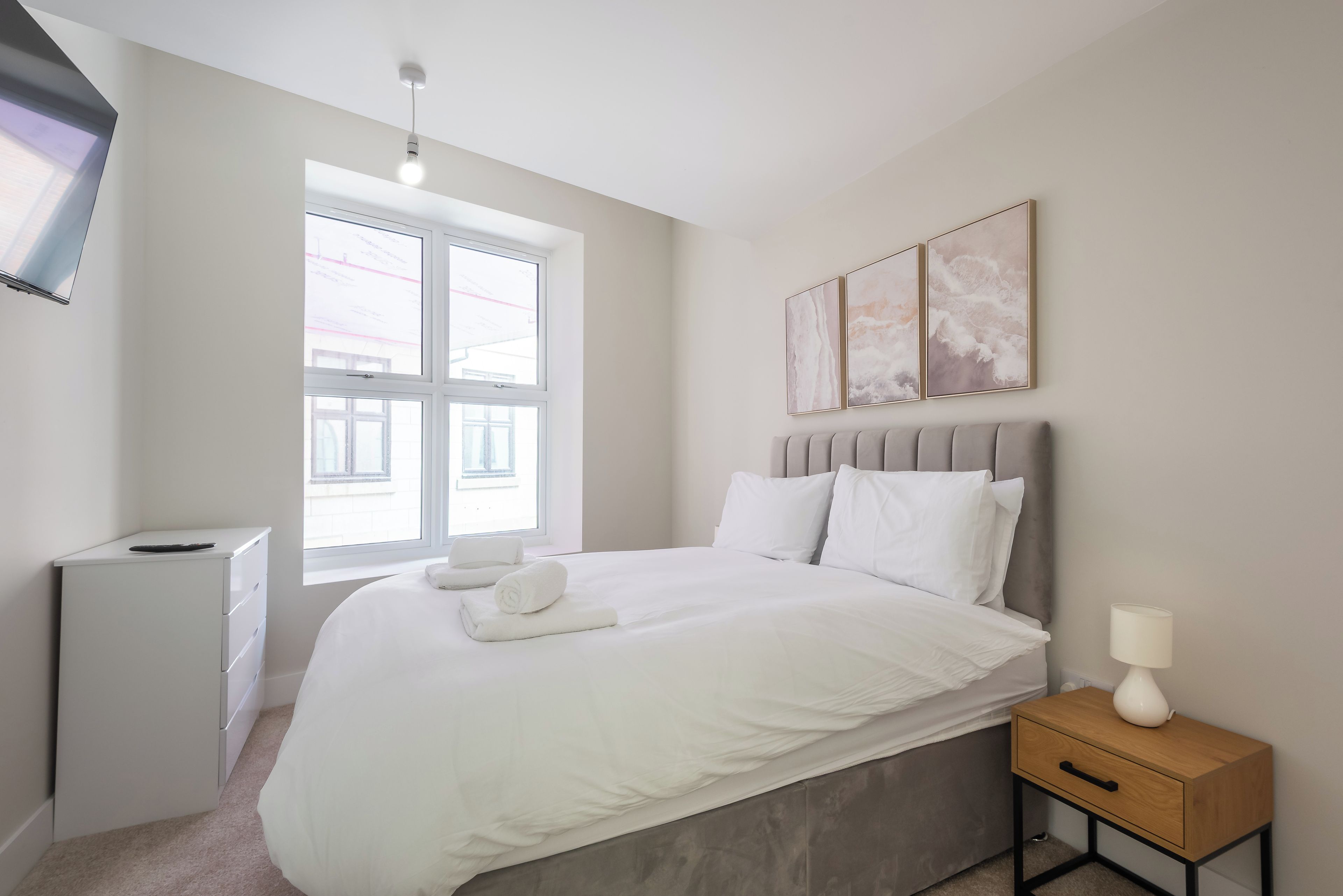 A bedroom with a bed, a bedside table, a lamp, a TV, and a window.