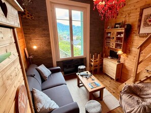 A cozy living room with a sofa, a coffee table, and a TV. There's a window with a view of greenery and mountains. - Apartment Near Mont-dore Centre (Mont-Dore)