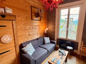 A cozy room with a wooden wall, a sofa, a coffee table, and a window with a view of a stone structure. - Apartment Near Mont-dore Centre (Mont-Dore)