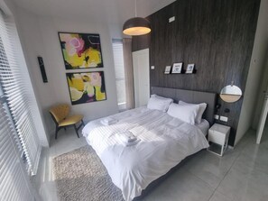 A modern bedroom with a large bed, a bedside table, a chair, and abstract artwork on the wall. - Jtowers G Royal Suites (Jerusalem)