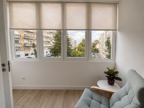 Ein Zimmer mit großem Fenster, grauem Sofa, kleinem runden Tisch und einem Blumentopf.
