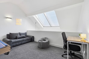 Una habitación moderna y compacta con techo inclinado, un escritorio integrado con lámpara, un sofá gris, un sillón de peluche y un cartel que dice 'LET'S STAY HOME'.