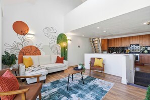 Een moderne woonkamer met een bank, fauteuils en een salontafel. In de achtergrond is een keuken zichtbaar met houten kastjes en een aanrecht.