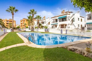 A swimming pool surrounded by well-maintained grass and palm trees, with a white building featuring a balcony in the background. - The Couthy Casa VillaMartin (Alicante)
