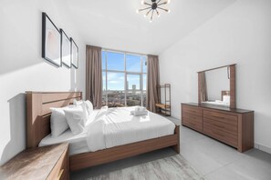 Quarto moderno com uma cama grande, cômoda de madeira, espelho e vista da cidade através de uma janela ampla.