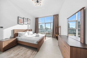 Ein modernes Schlafzimmer mit einem großen Bett, einem Nachttisch, einer Kommode und Blick auf die Stadt durch große Fenster.