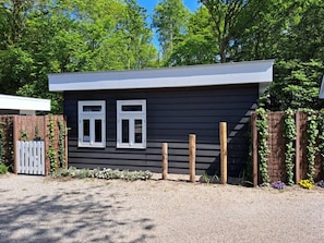 A small, dark-colored building with two windows, a gravel driveway, and a wooden fence with climbing plants. - Holiday Cottage With 1 Bedroom (Zoutelande)