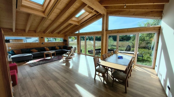 Quarto espaçoso com teto de madeira, sofá, mesa de jantar com cadeiras e grandes janelas com vista para o exterior.