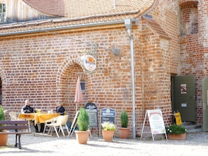 Ein Backsteinhaus mit einem Schild, auf dem 'Burgcafé in Spree' steht. Es gibt Tische und Stühle im Freien, Topfpflanzen und ein Schild für das Oktoberfest.