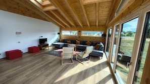 Ein modernes Wohnzimmer mit Holzbalkendecke, einem grauen Sofa, roten Hocker und einem Fernseher.