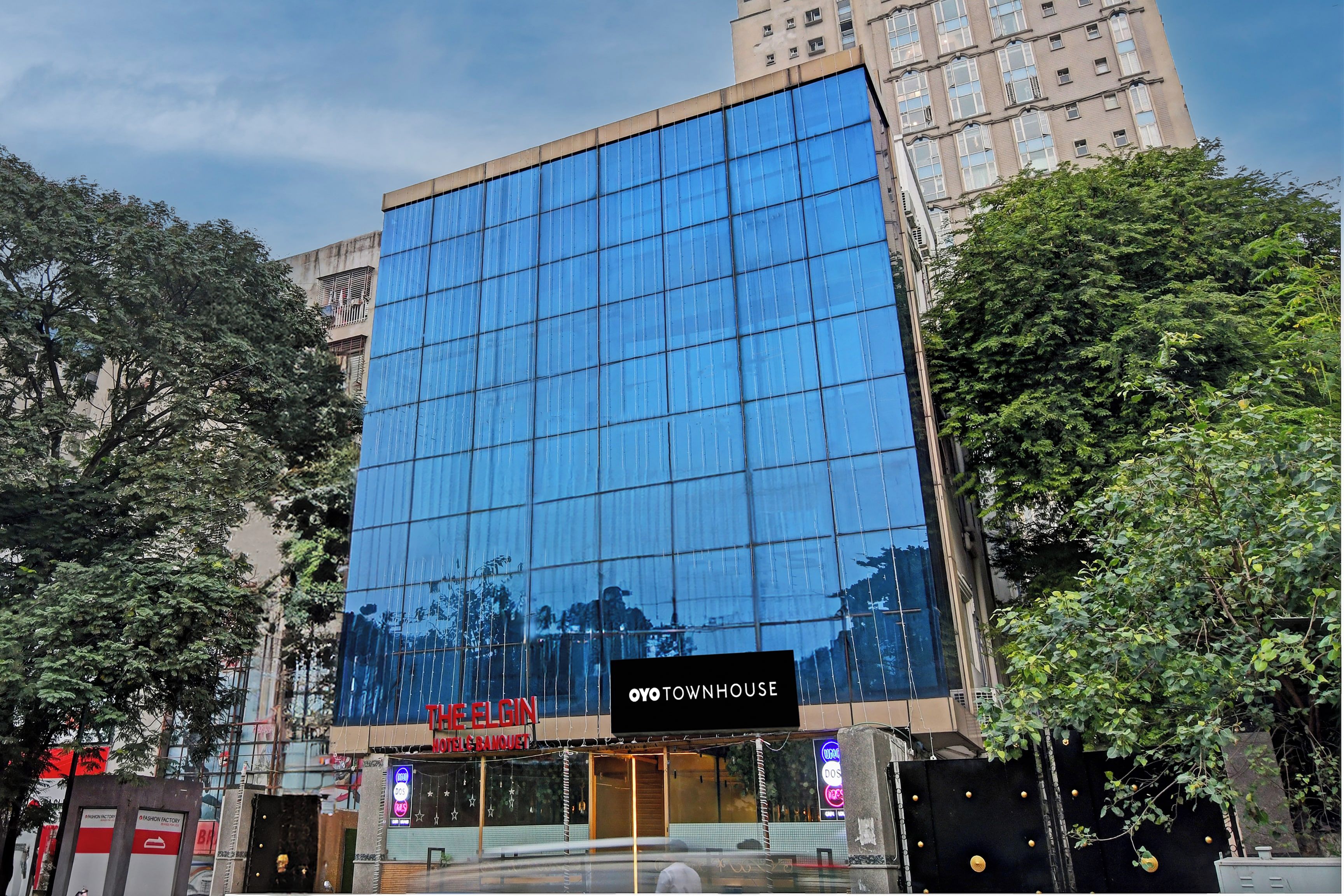 Un edificio moderno de hotel con fachada de vidrio, un cartel que dice 'The Elgin Hotel & Banquet' y un cartel que dice 'Oyo Townhouse'.