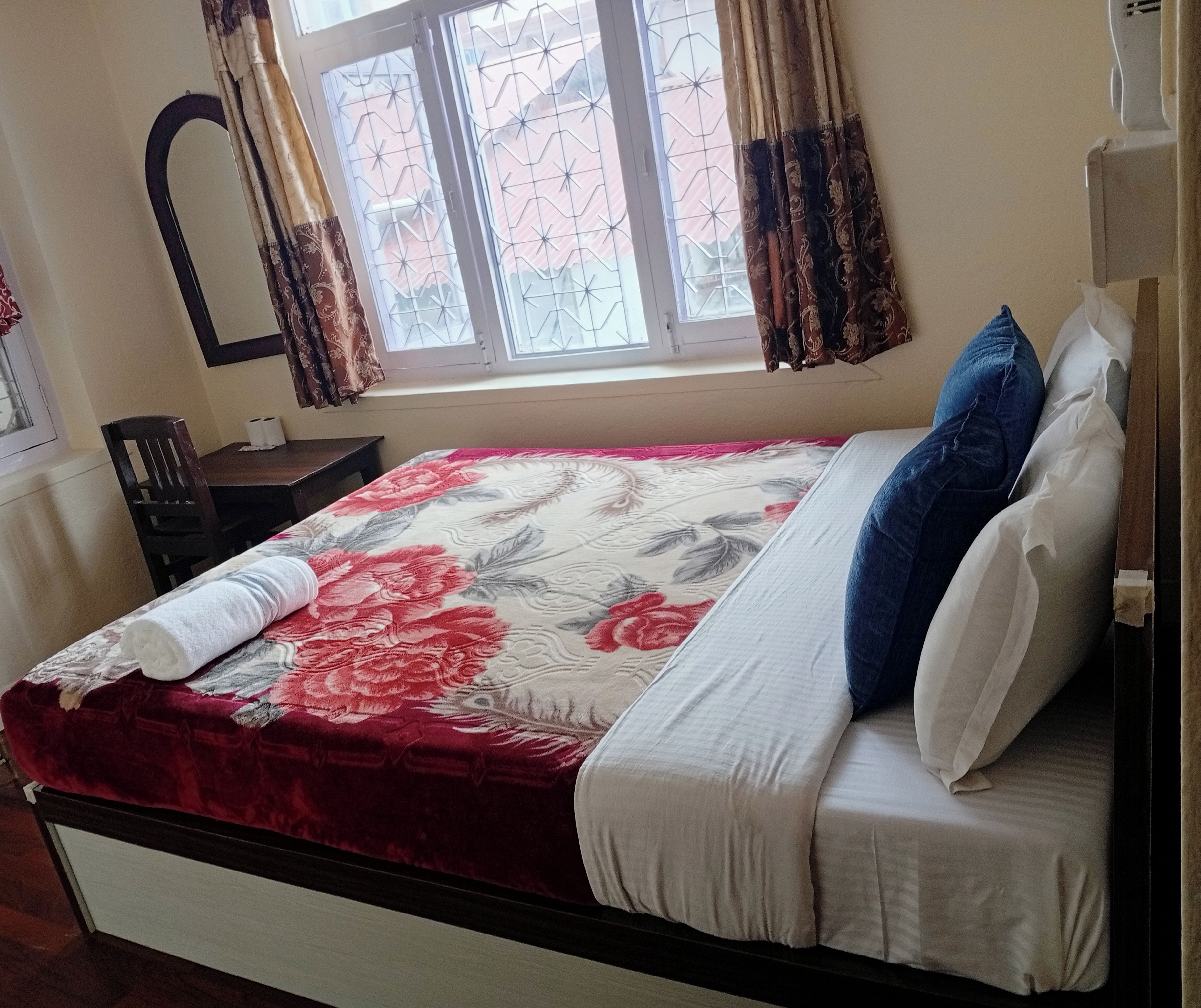 A bedroom with a bed, a nightstand, a chair, a window with curtains, and a wall-mounted mirror.