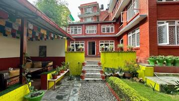 Une cour avec un bâtiment en briques rouges, un mur jaune et un petit espace aménagé avec un banc et des plantes en pot.