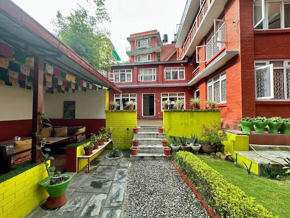 A courtyard with a red brick building, a yellow wall, and a small seating area with a bench and potted plants. - My Garden house (Kathmandu)