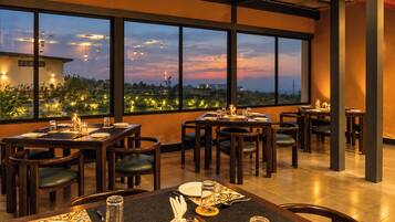 A restaurant with multiple tables and chairs, large windows offering a view of the cityscape at dusk.