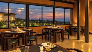 A restaurant with multiple tables and chairs, large windows offering a view of the cityscape at dusk. - Corbett KāSā (Ramnagar)