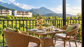 Un balcón con mobiliario de mimbre, un juego de té y un soporte escalonado para postres con pasteles, con vistas a un paisaje montañoso.
