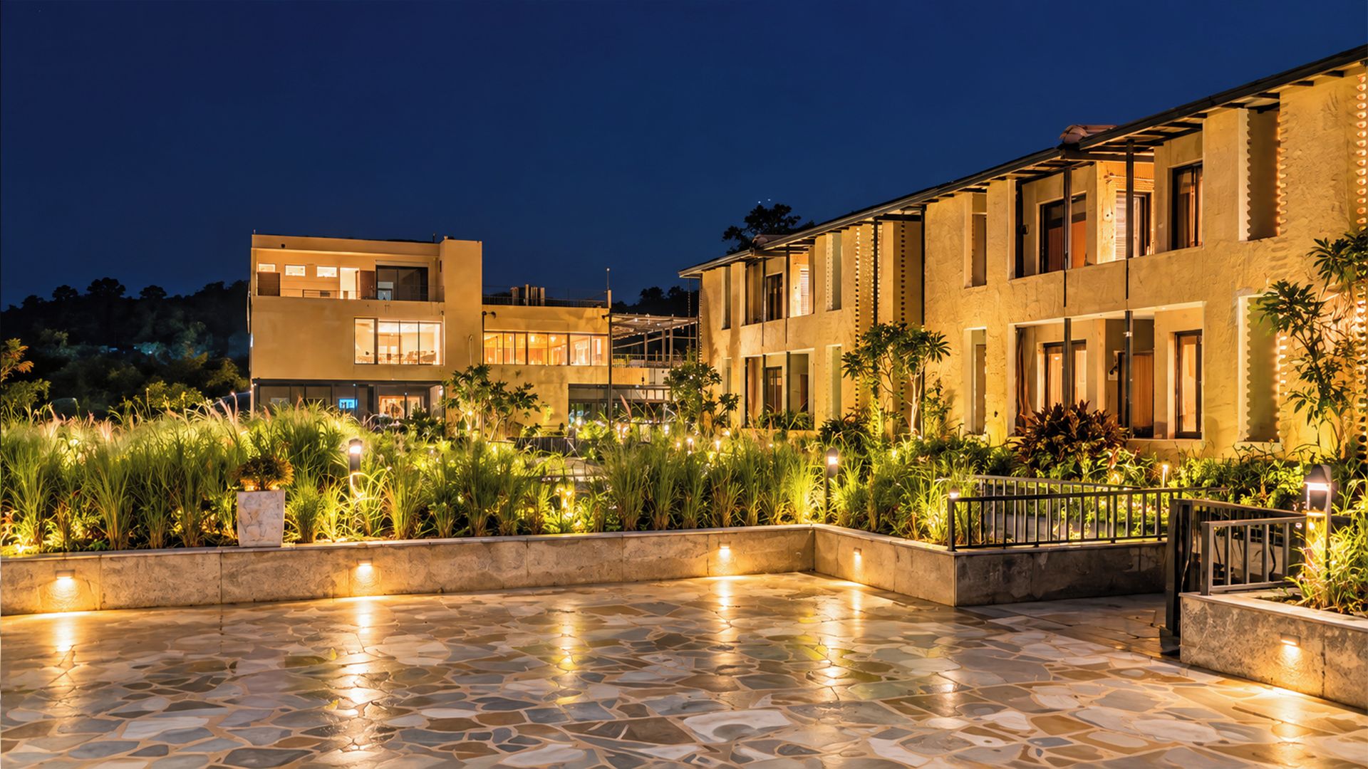 A courtyard with a stone floor, surrounded by buildings with large windows and outdoor lighting.