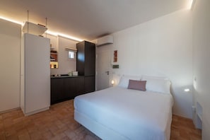 Quarto com cama, mesa de cabeceira, luminária na parede e porta que dá para uma área de cozinha.