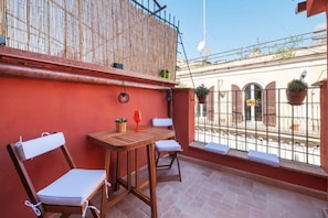 Varanda com mesa de madeira e duas cadeiras, parede vermelha e vista para um edifício com varandas.